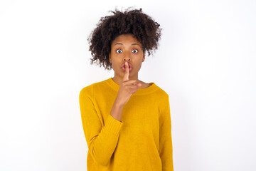 Wall Mural - Surprised young beautiful African American woman wearing yellow sweater against white wall makes silence gesture, keeps finger over lips and looks mysterious at camera