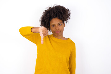 Wall Mural - young beautiful African American woman wearing yellow sweater against white wall looking unhappy and angry showing rejection and negative with thumbs down gesture. Bad expression.
