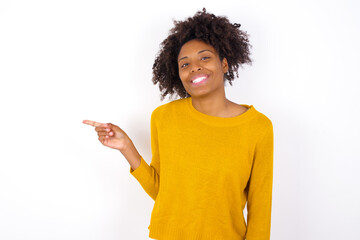 Wall Mural - young beautiful African American woman wearing yellow sweater against white laughs happily points away on blank space demonstrates shopping discount offer, excited by good news or unexpected sale.