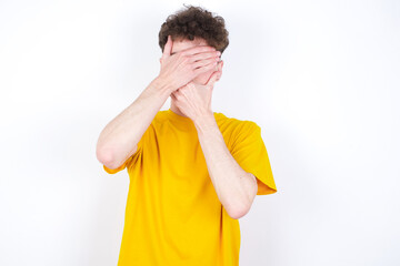 young caucasian handsome man with curly hair wearing yellow T-shirt against white studio background  Covering eyes and mouth with hands, surprised and shocked. Hiding emotions.