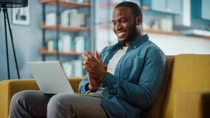 Aufkleber - Handsome Black African American Man Working from Home and Having Video Call on Laptop Computer while Sitting on a Sofa in Living Room. He is Clapping his Hands in Celebration of the Company's Success.
