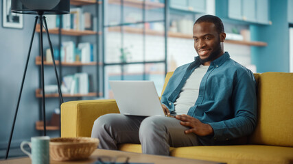 Wall Mural - Handsome Black African American Man Working on Laptop Computer while Sitting on a Sofa in Cozy Living Room. Freelancer Working From Home. Browsing Internet, Using Social Networks, Having Fun in Flat.