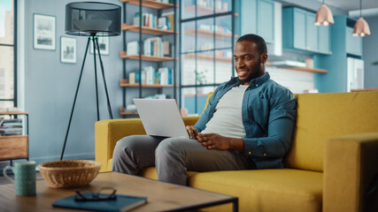 Aufkleber - Handsome Black African American Man Working on Laptop Computer while Sitting on a Sofa in Cozy Living Room. Freelancer Working From Home. Browsing Internet, Using Social Networks, Having Fun in Flat.
