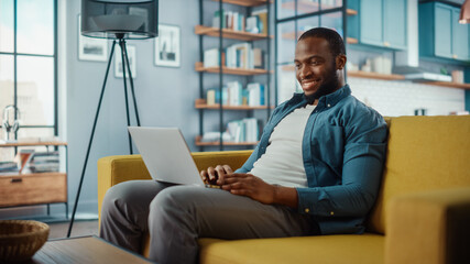 Aufkleber - Handsome Black African American Man Working on Laptop Computer while Sitting on a Sofa in Cozy Living Room. Freelancer Working From Home. Browsing Internet, Using Social Networks, Having Fun in Flat.