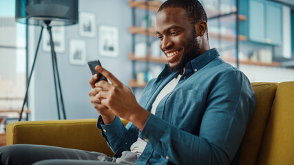 Aufkleber - Handsome Black African American Man Using Smartphone while Sitting on a Sofa in Cozy Living Room. Freelancer Working From Home. Browsing Internet, Using Social Networks, Having Fun in Flat.