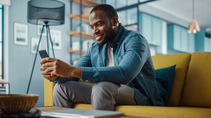 Aufkleber - Handsome Black African American Man Using Smartphone while Sitting on a Sofa in Cozy Living Room. Freelancer Working From Home. Browsing Internet, Using Social Networks, Having Fun in Flat.