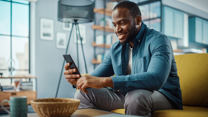 Aufkleber - Handsome Black African American Man Using Smartphone while Sitting on a Sofa in Cozy Living Room. Freelancer Working From Home. Browsing Internet, Using Social Networks, Having Fun in Flat.