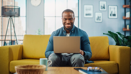 handsome black african american man working on laptop computer while sitting on a sofa in cozy livin