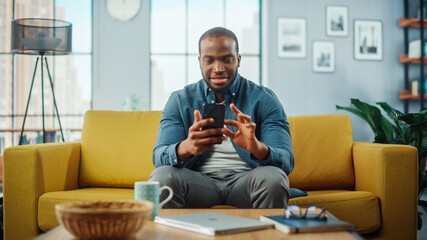 Aufkleber - Excited Black African American Man Using Smartphone while Resting on a Sofa in Living Room. Happy Man Smiling at Home and Chatting to Colleagues and Clients Over the Internet. Using Social Networks.