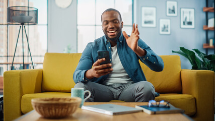 Aufkleber - Happy Black African American Man Having a Video Call on Smartphone while Sitting on a Sofa in Living Room. Excited Man Smiling at Home and Talking to Colleagues and Clients Over the Internet.