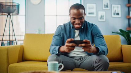 Aufkleber - Happy Black African American Man Having a Playing Video Game on Smartphone App while Sitting on a Sofa in Living Room. Excited Person of Color Resting at Home and Having Fun Over the Internet.