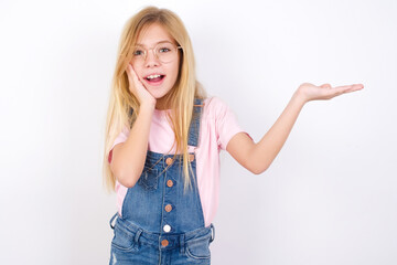 Wall Mural - Crazy beautiful caucasian little girl wearing jeans overall over white background advising discount prices hold open palm new product