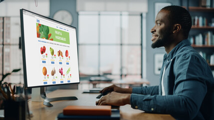 Wall Mural - Handsome Black African American is Using Desktop Computer with Groceries Delivery Web Page to Order Fresh Vegetables to Make Dinner. He Lives in Modern Huge Appatment.