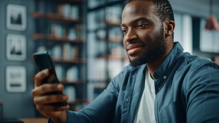 Aufkleber - Happy Black African American Man Using Smartphone while Sitting at Table in Living Room. Excited Man Smiling at Home and Talking to Colleagues, Clients and Friends Over the Internet.