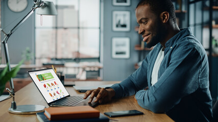 Wall Mural - Handsome Black African American is Using Laptop Computer with Groceries Delivery Web Page to Order Fresh Vegetables to Make Dinner. He Lives in Modern Huge Appatment.