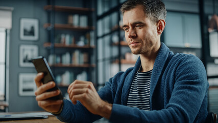 Aufkleber - Serious Caucasian Man Using Smartphone while Sitting at a Desk in Stylish Living Room. Young Man Smiling at Home and Chatting to Colleagues and Clients Over the Internet. Using Social Networks.