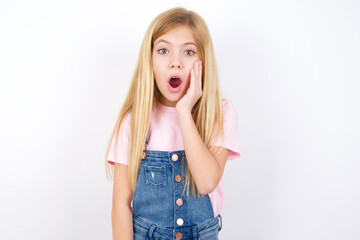 Wall Mural - Shocked beautiful caucasian little girl wearing denim jeans overall over white background looks with great surprisment being very stunned, astonished with unexpected news, Facial expressions concept.