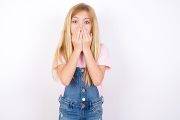 Wall Mural - Vivacious beautiful caucasian little girl wearing denim jeans overall over white background , giggles joyfully, covers mouth, has natural laughter, hears positive story or funny anecdote