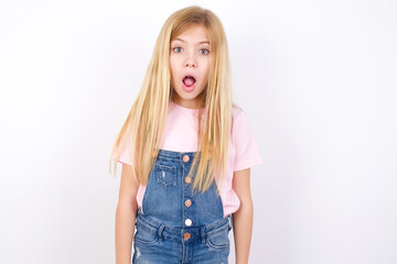 Wall Mural - Oh my God. Surprised beautiful caucasian little girl wearing denim jeans overall over white background stares at camera with shocked expression exclaims with unexpectedness,