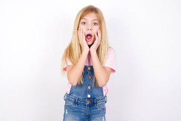 Scared terrified beautiful caucasian little girl wearing denim jeans overall over white background shocked with prices at shop, People and human emotions concept