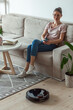 © perfectlab - Robotic vacuum cleaner cleaning carpet, woman remote control mobile phone and enjoy rest, sitting on sofa home