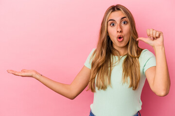 Young blonde caucasian woman isolated on pink background holds copy space on a palm, keep hand over cheek. Amazed and delighted.