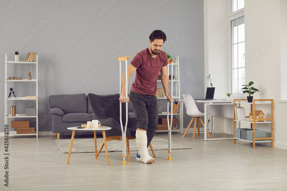 Rehabilitation and recovery of people after physical injury such as bone fracture in car accident: Man with broken leg in plaster cast stands up from chair and starts walking with crutches at home