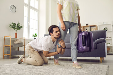 Young married couple breaking up. Angry woman leaving home with packed suitcase. Clingy desperate husband on floor holding wife's leg begging his love to stay. Relationship breakup and divorce concept