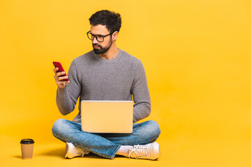 Young bearded man sitting on the floor with laptop and talking at phone. Isolated over yellow background.