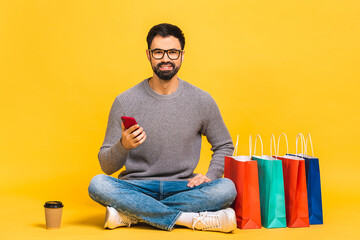 Online home shopping time! Portrait of young bearded man sitting on the floor with mobile phone with crossed legs with shopping bags.