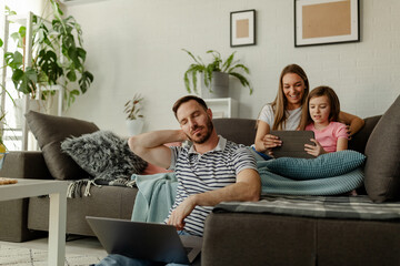 Wall Mural - Young mother and girl are playing games, smiling while dad is sitting at floor, looks so tired.