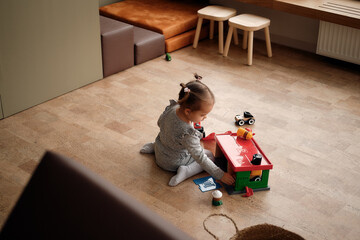 Little girl in a gray dress plays in the children's room with a set of garage and cars. Stylish and modern children's room. View from above.