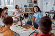 © Ivan - Young tutor is teaching to read her student. Elementary school kids sitting on desks and reading books in classroom