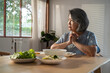 © Kawee - Asian senior woman eating food, sitting alone on dining table at home.