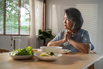 Asian senior woman eating food, sitting alone on dining table at home.