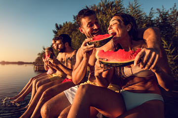 Friends sitting on the edge of a pier enjoying on a summer day at the lake eating watermelon.	