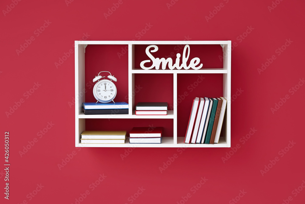 Shelf with books, alarm clock and decor hanging on color wall