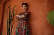 Portrait of beautiful dark-haired woman with crown braid hairstyle and floral head band wearing chic chiffon dress with basque belt standing at the orange ethnic wall among cactuses. Mexican style