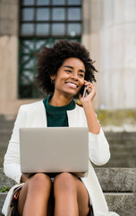 Wall Mural - Business woman talking on phone and using laptop outdoors.