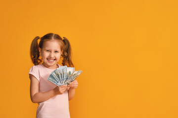 cute smiling little girl with two ponytails holds a wad of dollar bills on a yellow-orange background in the studio. The concept of winning, depositing and investing