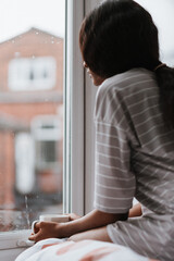 Young African female with a cup of tea looking out of a window while sitting on the bed