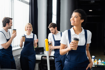 Wall Mural - African american cleaner with takeaway drink standing near colleagues talking in office