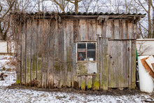 An Old Wooden Utility Shed With Rotten Planks. Early Spring With Remnants Of Snow On The Ground.