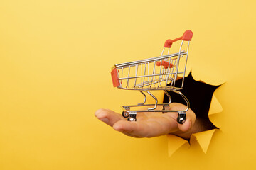 Hand through a hole with a mini grocery shopping trolley on a yellow paper background. Sales or shopping concept