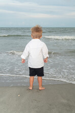 Little Boy In Black Shorts And White Shirt Looks Out At The Ocean