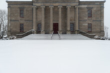 A Vintage Government Building With Large Round Pillars, Red Wooden Doors, Black Metal Rails, Grey Marble Steps And Brick Entrance. It Is Snowing Heavily And The Large Steps Are Covered In White Snow. 