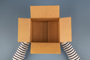 Wall Mural - Female hands with empty open cardboard box on gray background. Top view