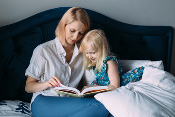 Happy family of blonde mom and daughter are reading a book in bed.