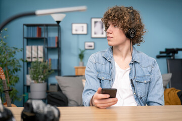 Wall Mural - Portrait of Teenager student learning music sitting at desk in his living room at home. The boy wearing blue shirt has headphones on head and holding a smartphone looking window.