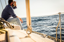 Man Sit Down On Sail Boat Deck And Look Far At The Ocean - Adult Male People Enjoy Travel And Lifestyle - Marine And Navigation Concept Image - Coast In Background - Summer Excursion
